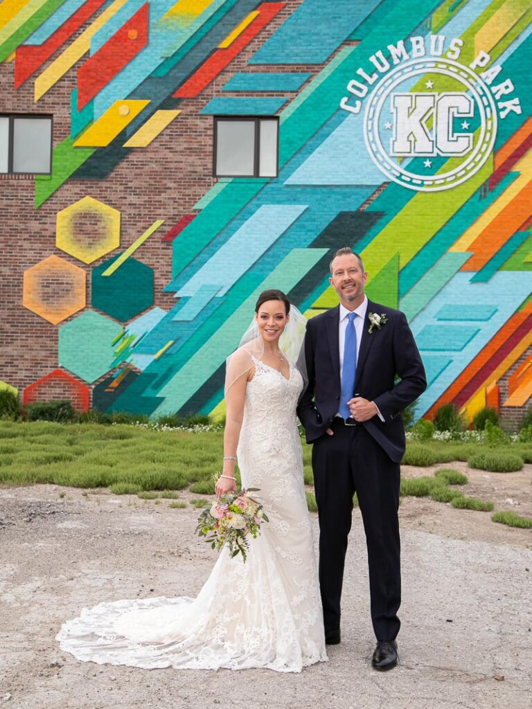 The bride and groom smiling in front of a colorful brick wall